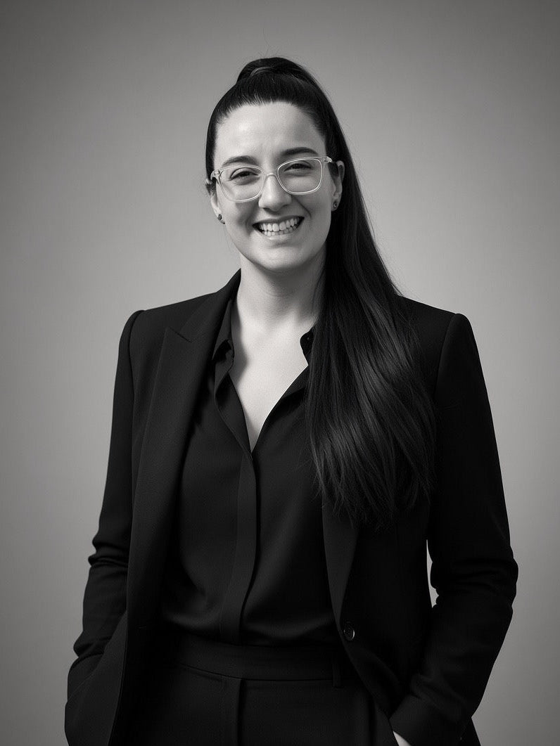 A black and white portrait of a woman with long dark hair, wearing a black blazer and matching top, standing against a neutral gray background.