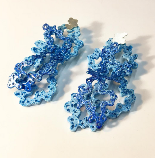 A pair of dangling statement earrings with a glittery, coral-like organic shape in shades of light and dark blue, photographed on a plain off-white background.